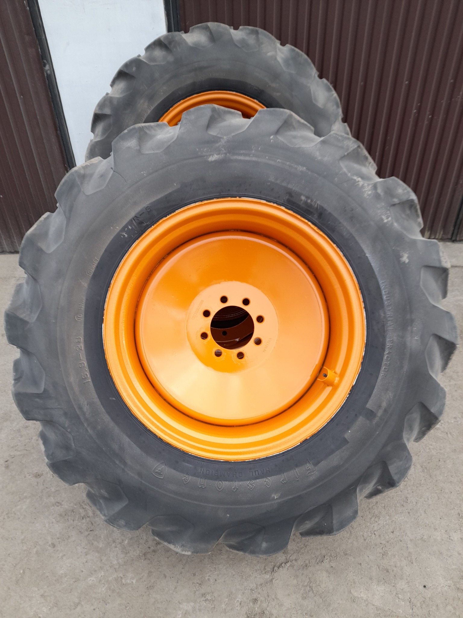 Dwa duże, czarne opony ciągnikowe z pomarańczowymi felgami, ułożone jeden za drugim na betonowej powierzchni, na tle brązowej, metalowej ściany.