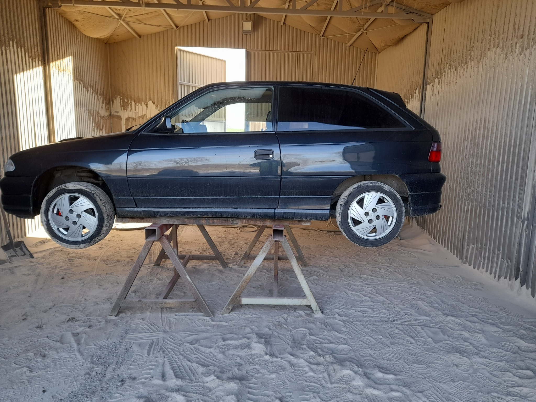 Czarny samochód osobowy, podniesiony na metalowych kozłach w warsztacie, przygotowywany do prac blacharsko-lakierniczych. Podłoga i ściany pokryte pyłem.