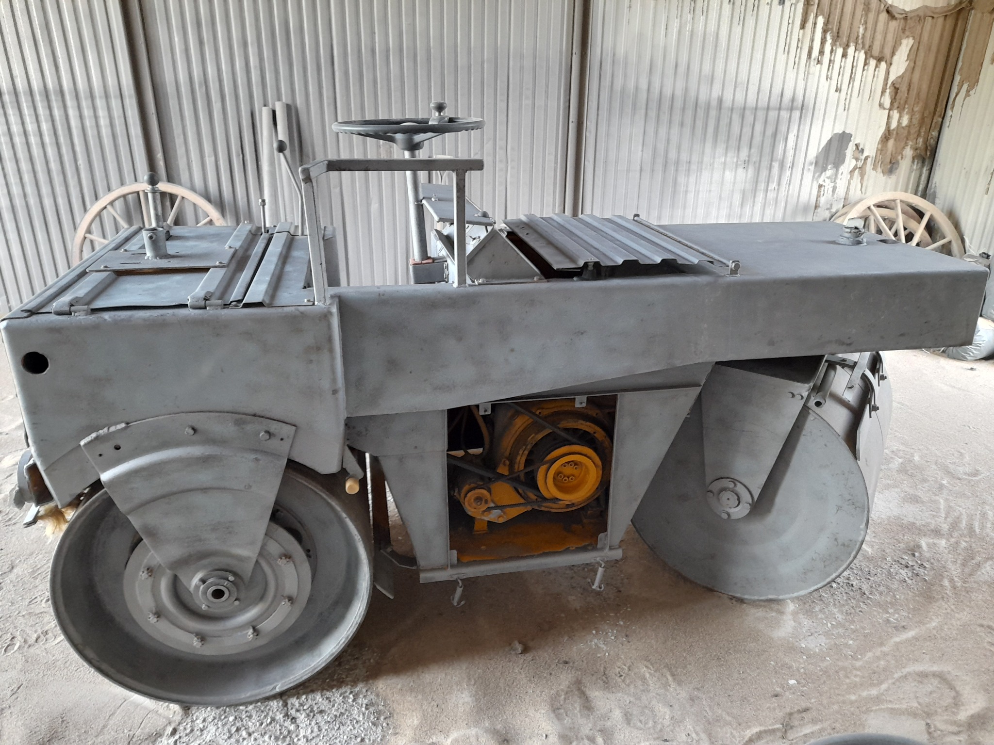 Szary walec drogowy po renowacji, z widocznym żółtym silnikiem i drewnianymi kołami w tle.