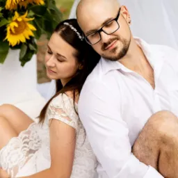 Para młoda siedzi plecami do siebie, ona w koronkowej sukience i ozdobą we włosach, on w białej koszuli, obok bukiet słonecznik&oacute;w, tło rozmyte.