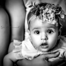 Portret niemowlęcia z dużymi oczami, w czarno-białej tonacji, z ozdobną opaską z kwiatami na głowie, trzymanego na rękach.