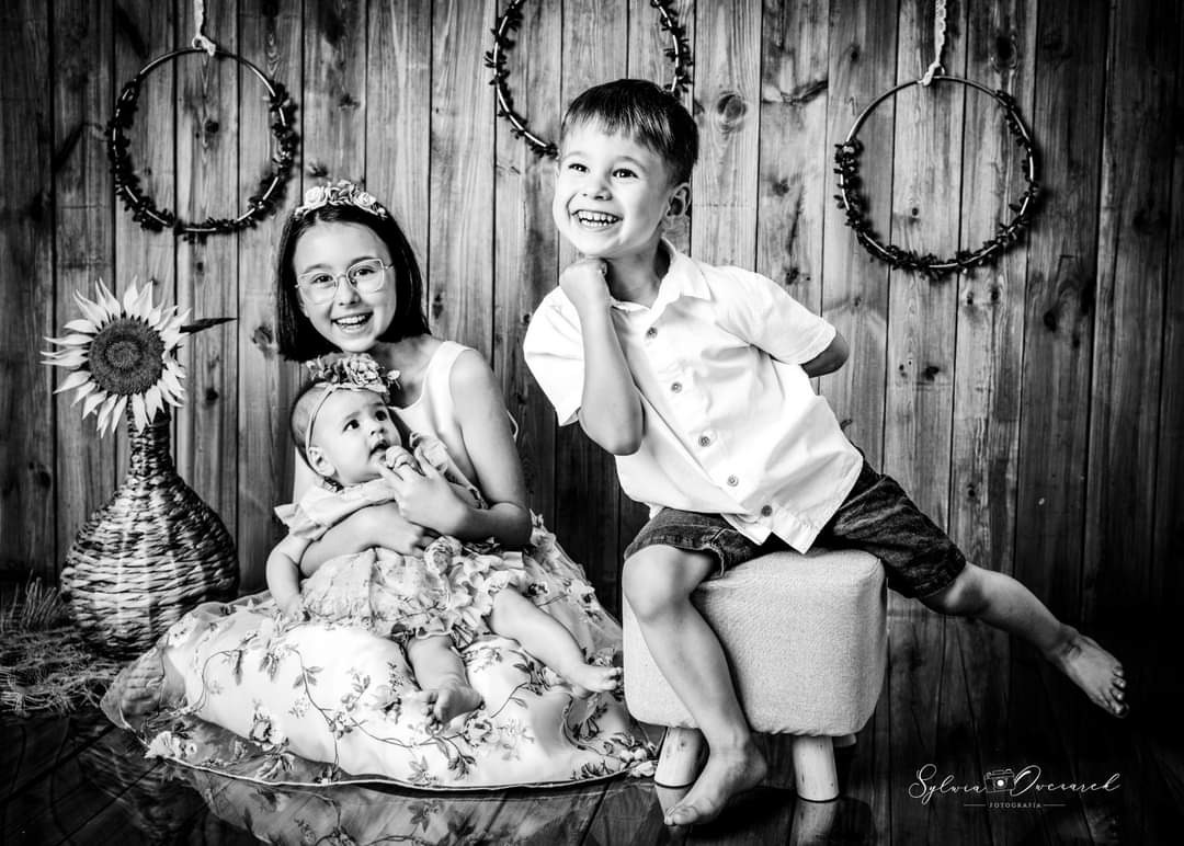 Czarno-białe zdjęcie studyjne przedstawia uśmiechniętą dziewczynkę w okularach trzymającą na kolanach niemowlę w kwiecistej sukience, obok siedzi roześmiany chłopiec na taborecie, w tle drewniana...