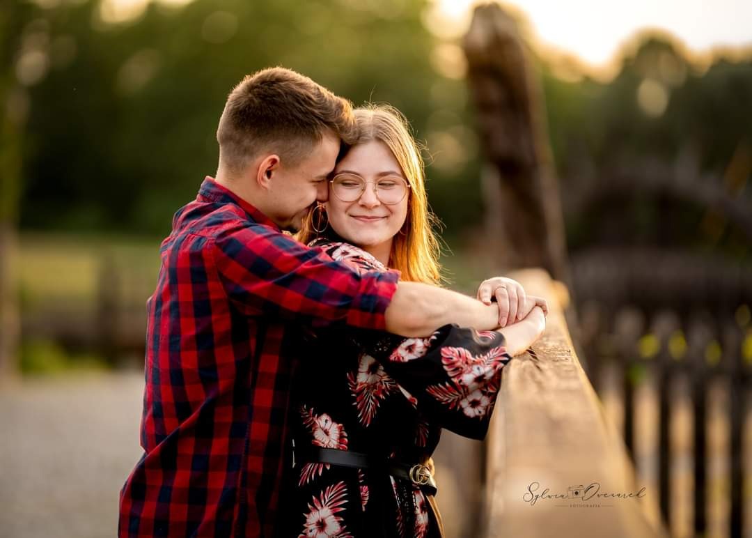 Młoda para w objęciach na drewnianym moście w promieniach zachodzącego słońca, on w czerwono-czarnej koszuli w kratę, ona w kwiecistej sukience i okularach, delikatny uśmiech.
