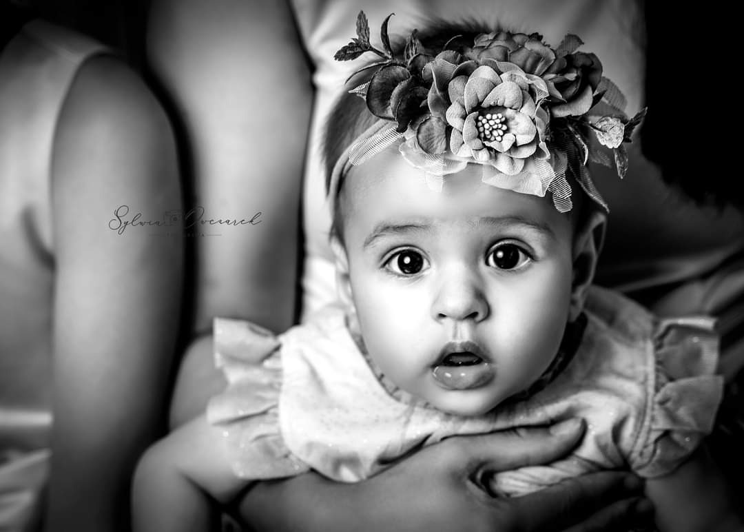 Portret niemowlęcia z dużymi oczami, w czarno-białej tonacji, z ozdobną opaską z kwiatami na głowie, trzymanego na rękach.