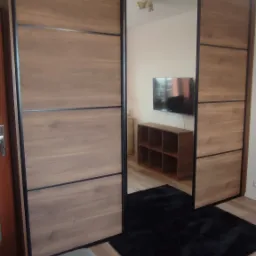 Duża szafa wnękowa z przesuwnymi drzwiami z lustrem, z drewnianymi panelami i czarnymi ramami, widoczna w tle telewizor i regał.