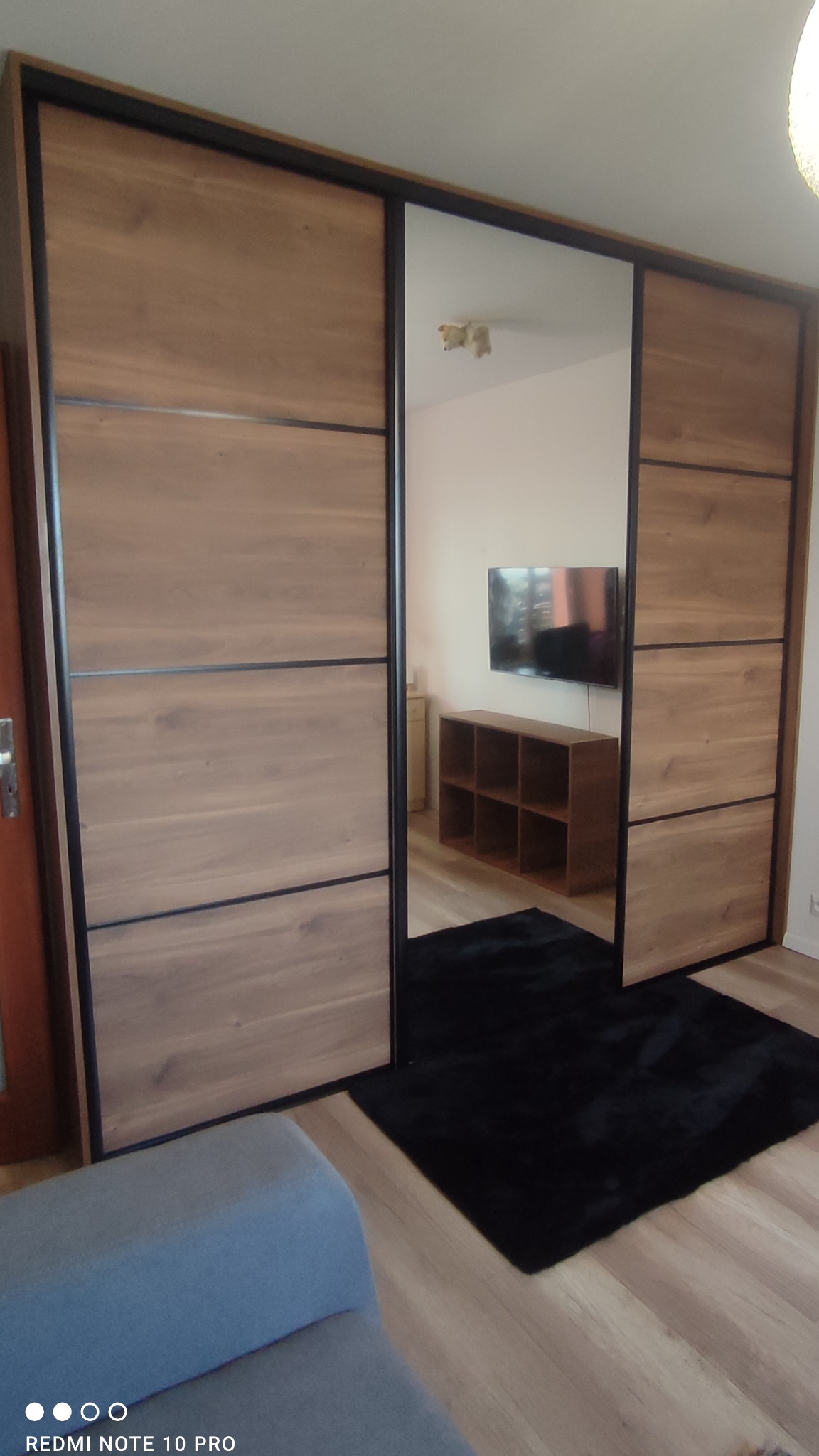 Duża szafa wnękowa z przesuwnymi drzwiami z lustrem, z drewnianymi panelami i czarnymi ramami, widoczna w tle telewizor i regał.