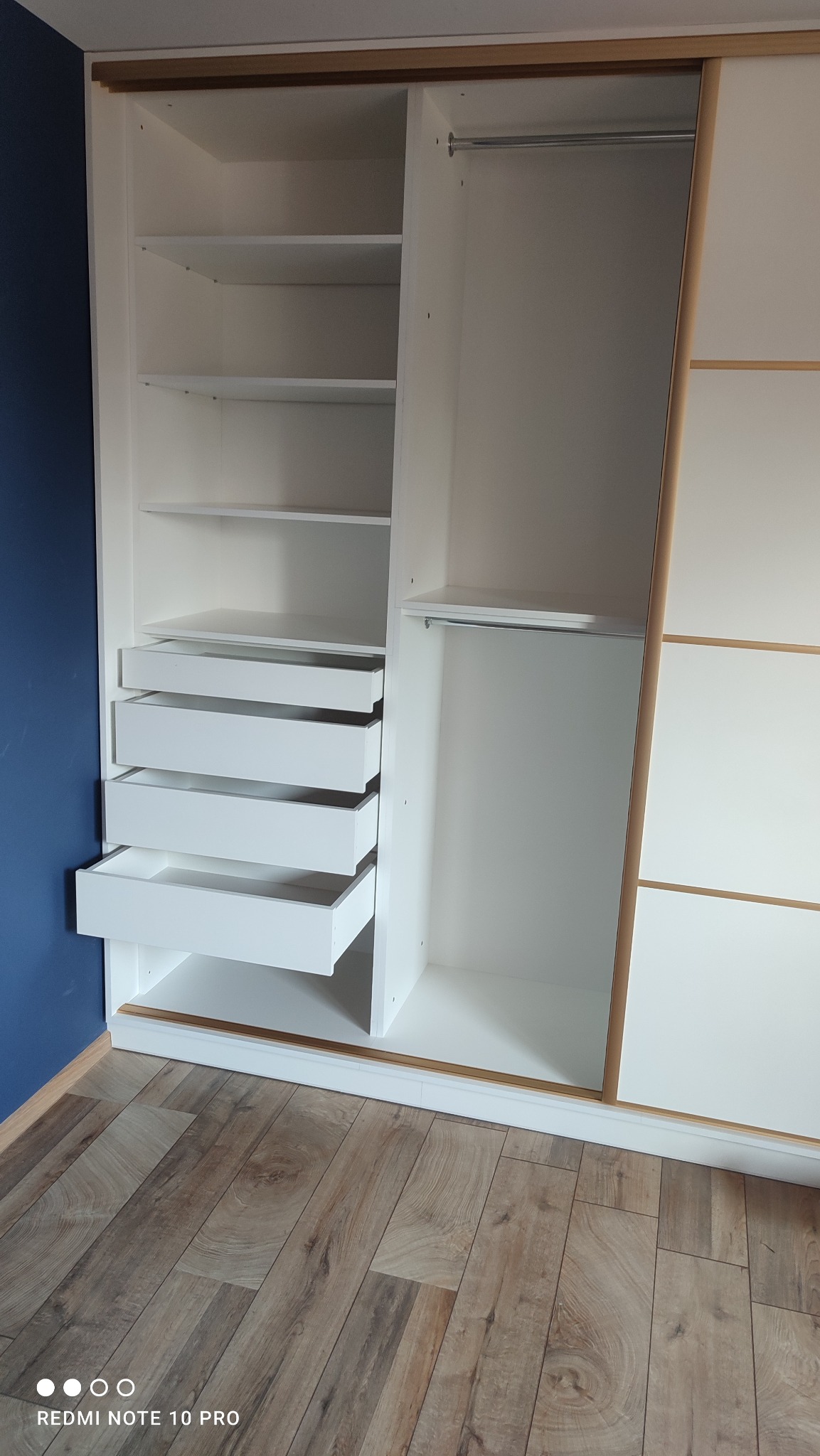 Biała szafa przesuwna z otwartymi szufladami, widoczne półki i drążek na ubrania, wykończenie z jasnego drewna, na podłodze panele, ściana pomalowana na niebiesko.