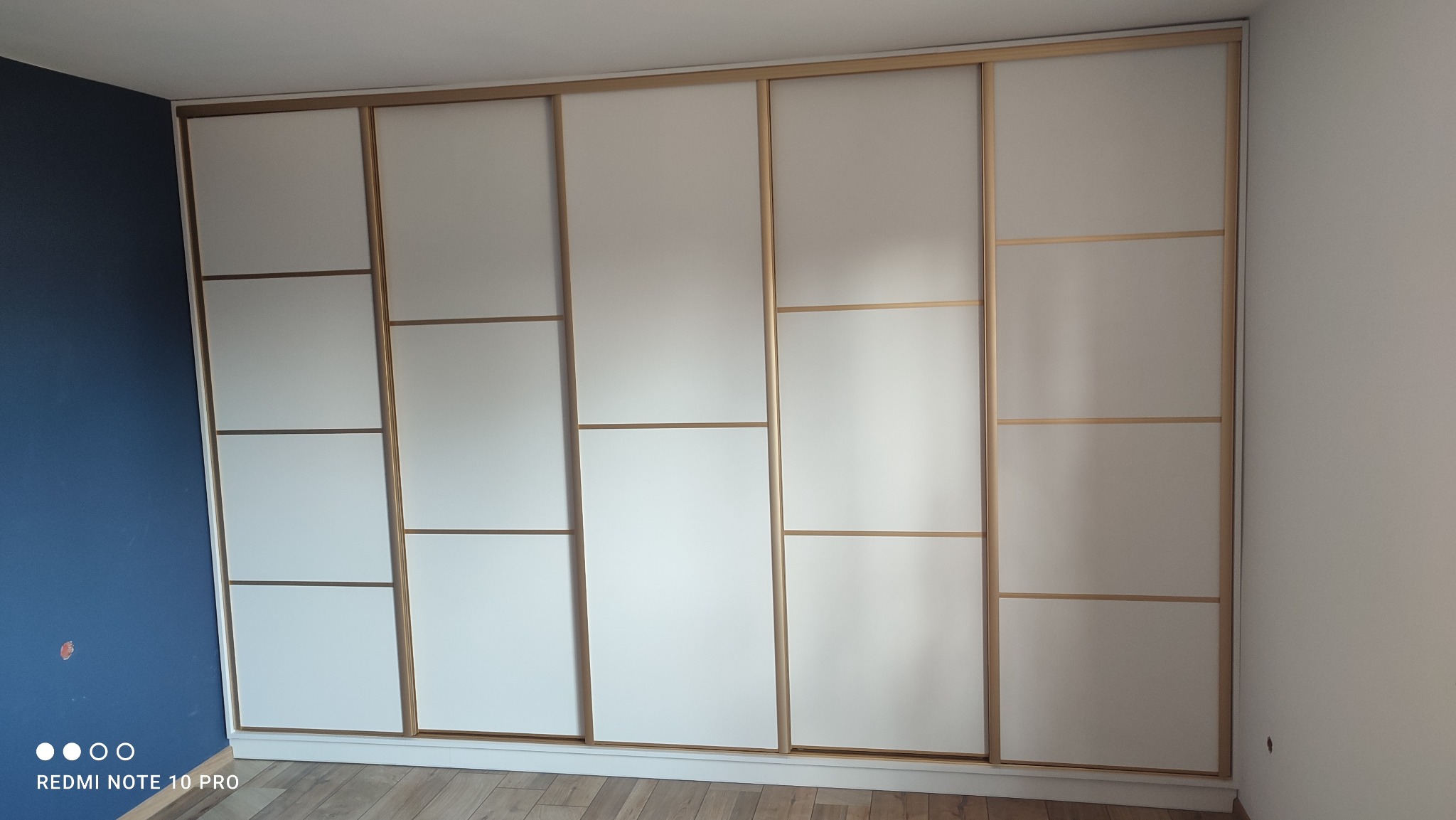 Biała szafa wnękowa z przesuwnymi drzwiami, podzielona poziomymi listwami w kolorze jasnego drewna, widoczna na tle błękitnej ściany.