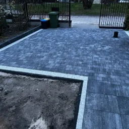 Ułożona szara kostka brukowa z jasną obwódką, widoczne śmietniki w tle i brama wjazdowa, częściowo odsłonięty teren z ziemią po lewej stronie.