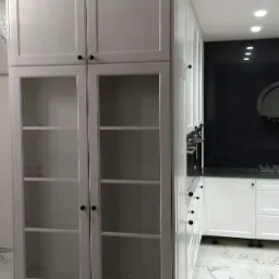 Biała, wysoka szafa z przeszklonymi drzwiami i półkami, część zabudowy kuchennej z białymi frontami i czarnym panelem na ścianie, marmurowa podłoga.