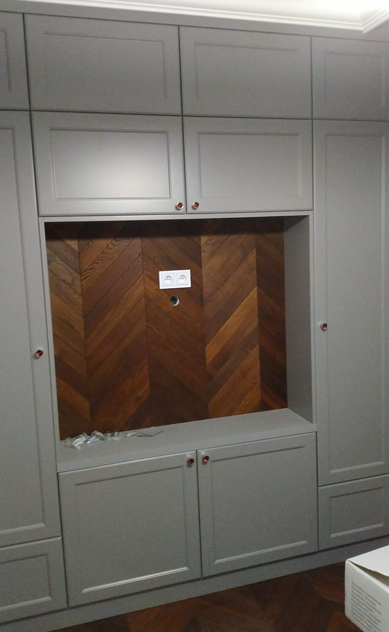 Szara zabudowa meblowa z drewnianą ścianą w jodełkę, widoczne gniazdka elektryczne i elementy montażowe na półce