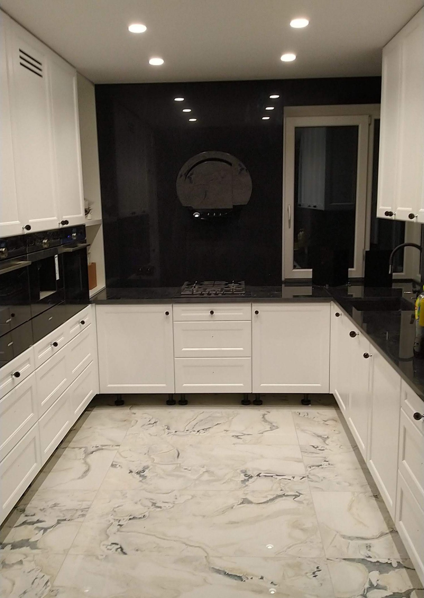 Wnętrze kuchni z białymi szafkami, czarnym blatem i marmurową podłogą, oświetlone punktowymi lampami sufitowymi, z widoczną płytą gazową i czarnym panelem na ścianie.