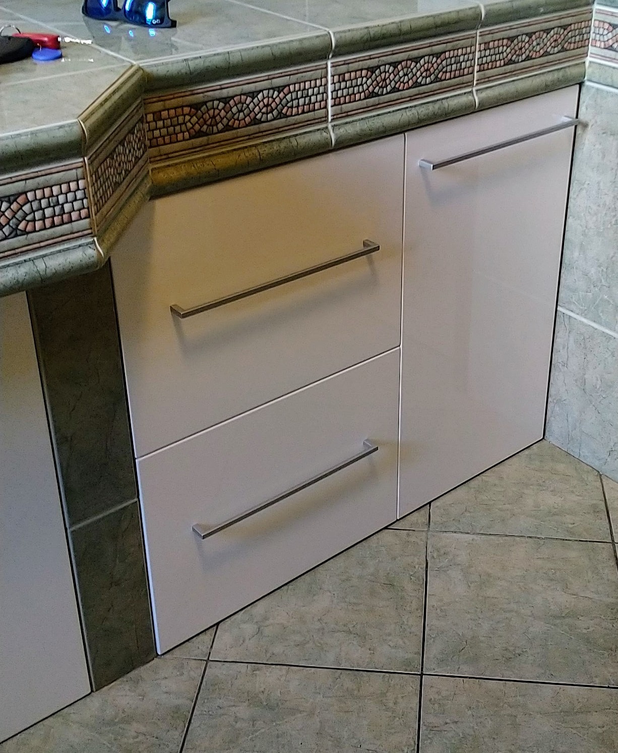 Biała szafka kuchenna z dwoma szufladami i drzwiczkami, z poziomymi, metalowymi uchwytami, widoczna spod kątem, z blatem wykończonym dekoracyjnymi płytkami ceramicznymi w odcieniach zieleni i brązu...