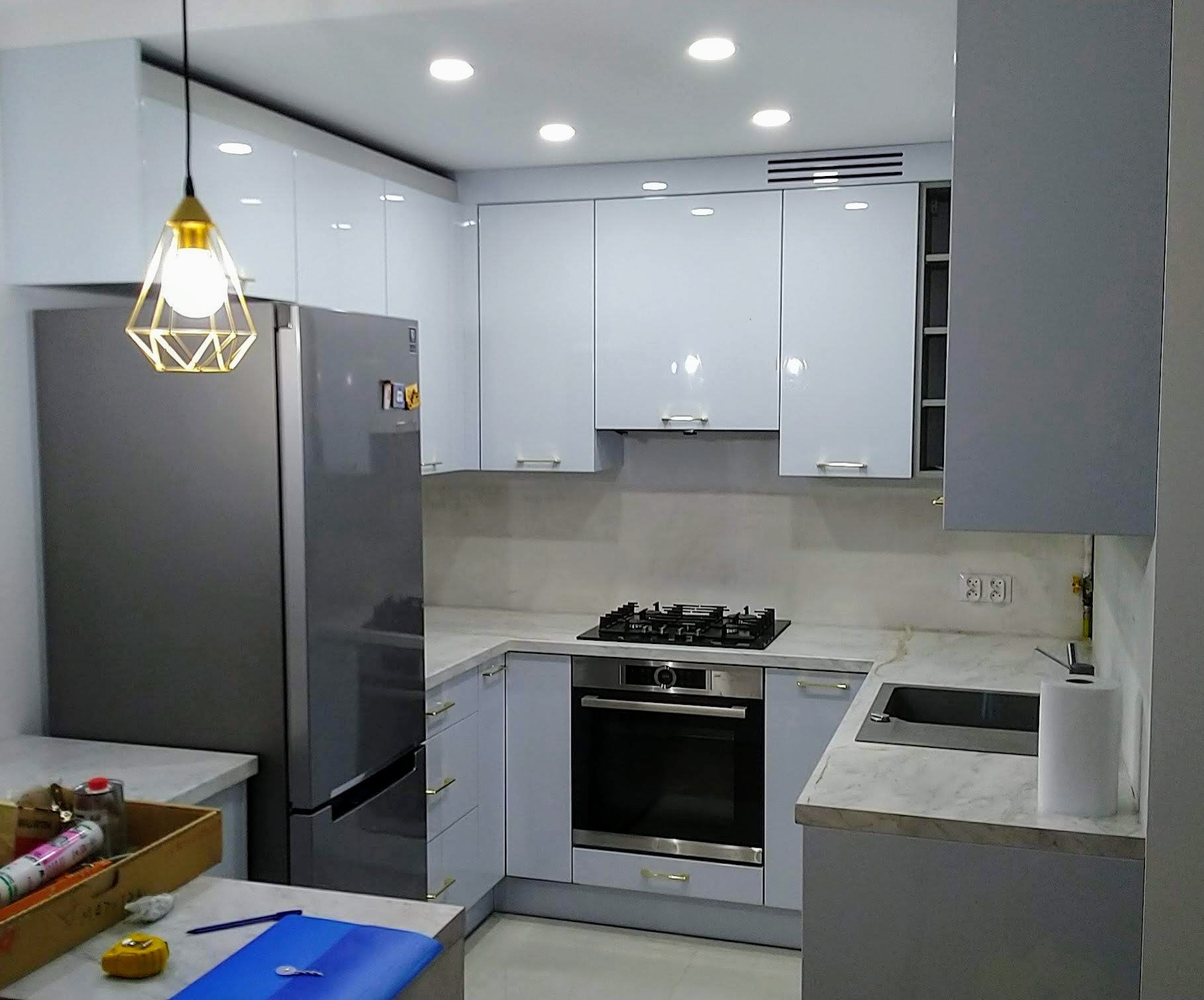 Nowoczesna kuchnia z białymi szafkami, szarym blatem imitującym marmur, srebrną lodówką i geometryczną lampą wiszącą z elementami w kolorze złota.