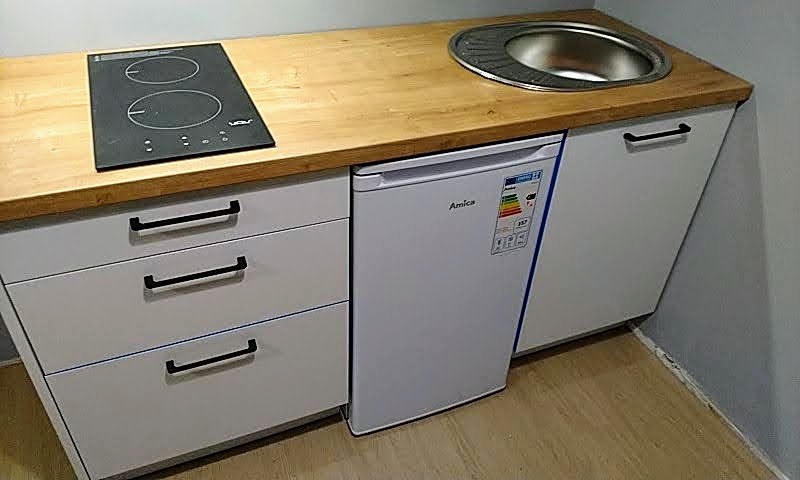 Kompaktowa zabudowa kuchenna z drewnianym blatem, płytą indukcyjną, zlewozmywakiem i małą lodówką Amica, fronty szafek w kolorze jasnoszarym z czarnymi uchwytami.