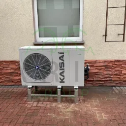 Zewnętrzna jednostka klimatyzacji Kaisai zamontowana na metalowej konstrukcji przed budynkiem z ceglaną podmurówką i oknem na górze.