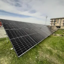 Instalacja paneli słonecznych na trawie przed budynkiem, widoczne metalowe ramy i moduły fotowoltaiczne ułożone rzędami, pochmurne niebo w tle.