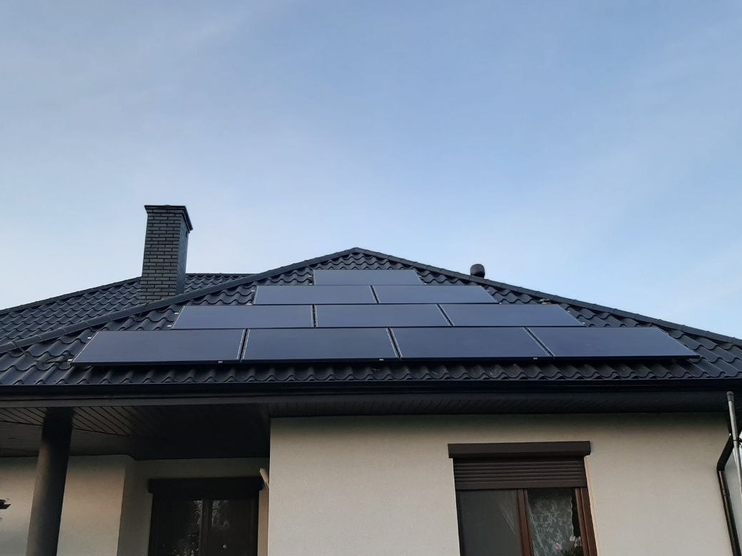 Czarny dach z panelami słonecznymi ułożonymi w piramidę, komin z cegły wystający ponad dach, biała elewacja domu z oknem z roletami.