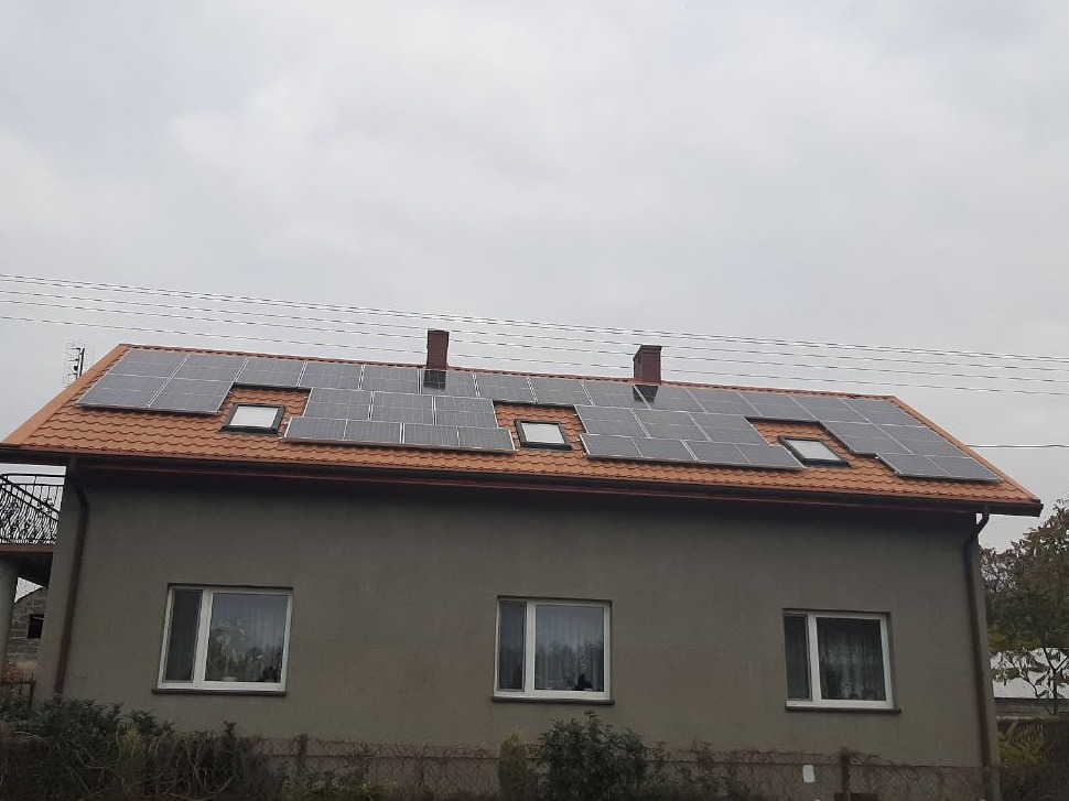 Dom jednorodzinny z panelami słonecznymi na pomarańczowym dachu, widoczne okna dachowe i kominy, pochmurne niebo w tle.