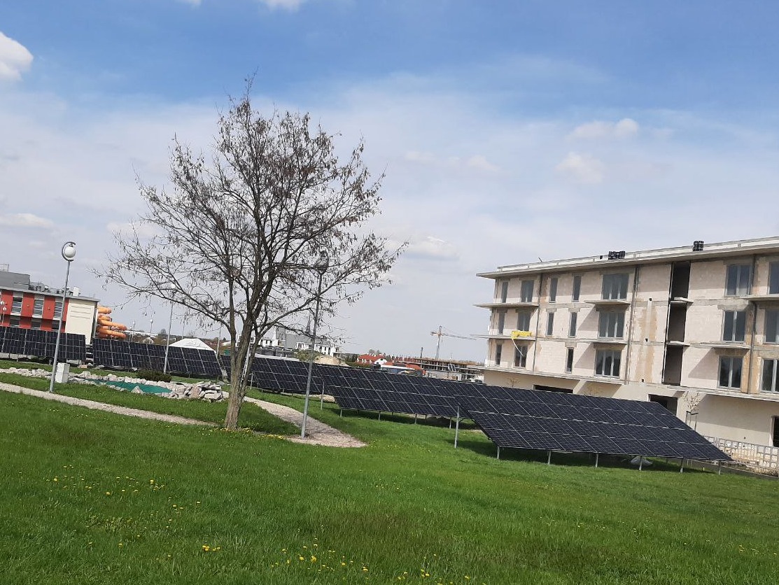 Rząd paneli fotowoltaicznych na zielonym trawniku przed budynkiem w trakcie budowy, w tle drzewo bez liści i błękitne niebo z chmurami.