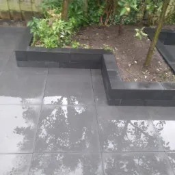 Szare, kwadratowe płyty tarasowe ułożone wokół podwyższonych rabat z ciemnoszarych bloczków, odbijające w deszczu korony drzew