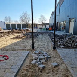 Plac budowy z częściowo ułożoną szarą kostką brukową, widoczne przygotowane podłoże z piasku, stosy kostki i narzędzia pomiarowe w tle, budynek z czarną elewacją.