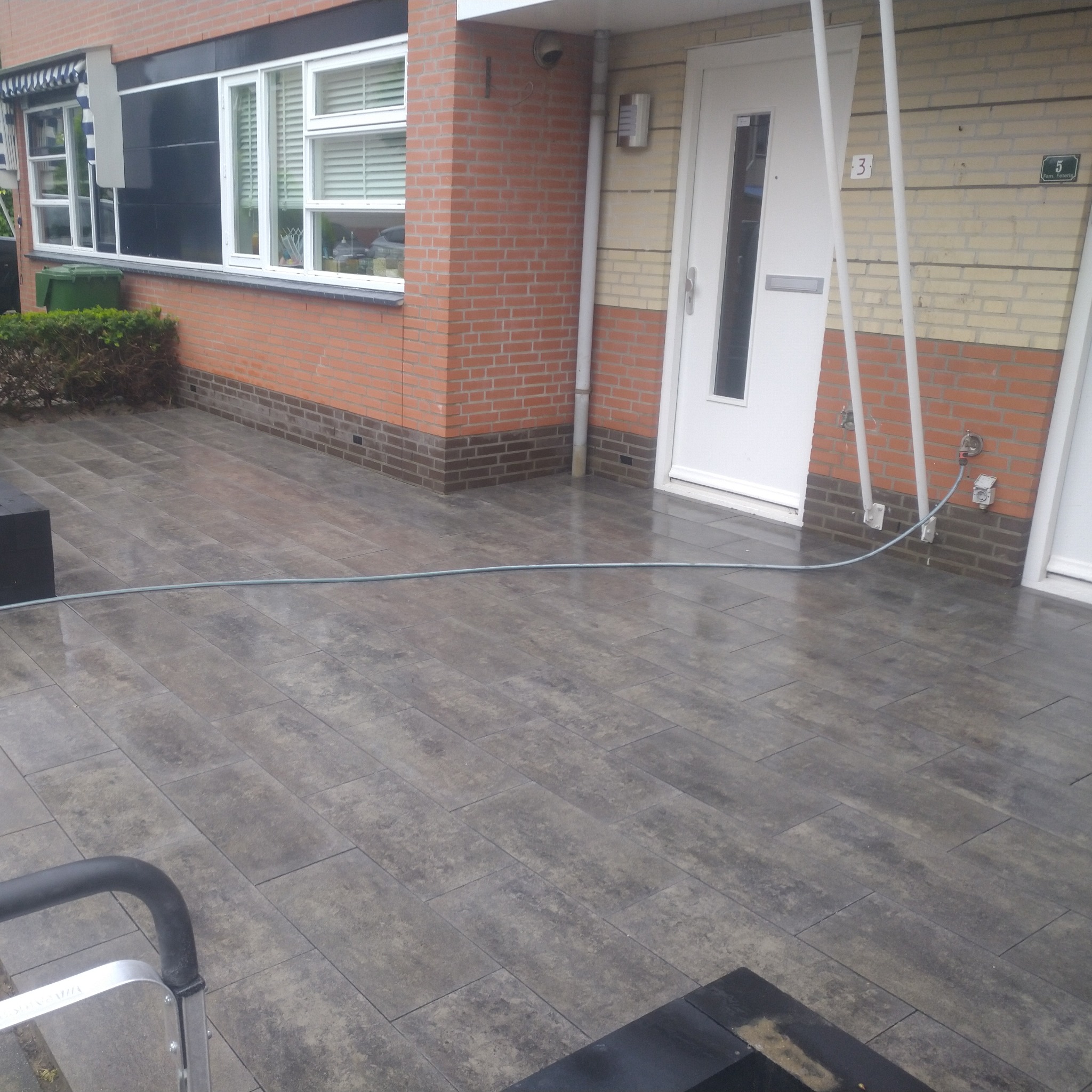 Szary, prostokątny chodnik z kostki brukowej prowadzący do drzwi wejściowych domu z cegły, widoczny wąż ogrodowy i fragment drabiny.