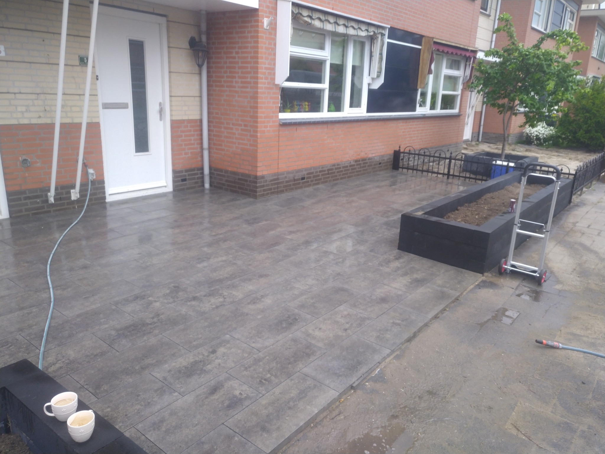 Nowo ułożona, mokra kostka brukowa w odcieniach szarości przed domem z cegły, z dwoma filiżankami kawy na niskim murku i wężem ogrodowym w tle.