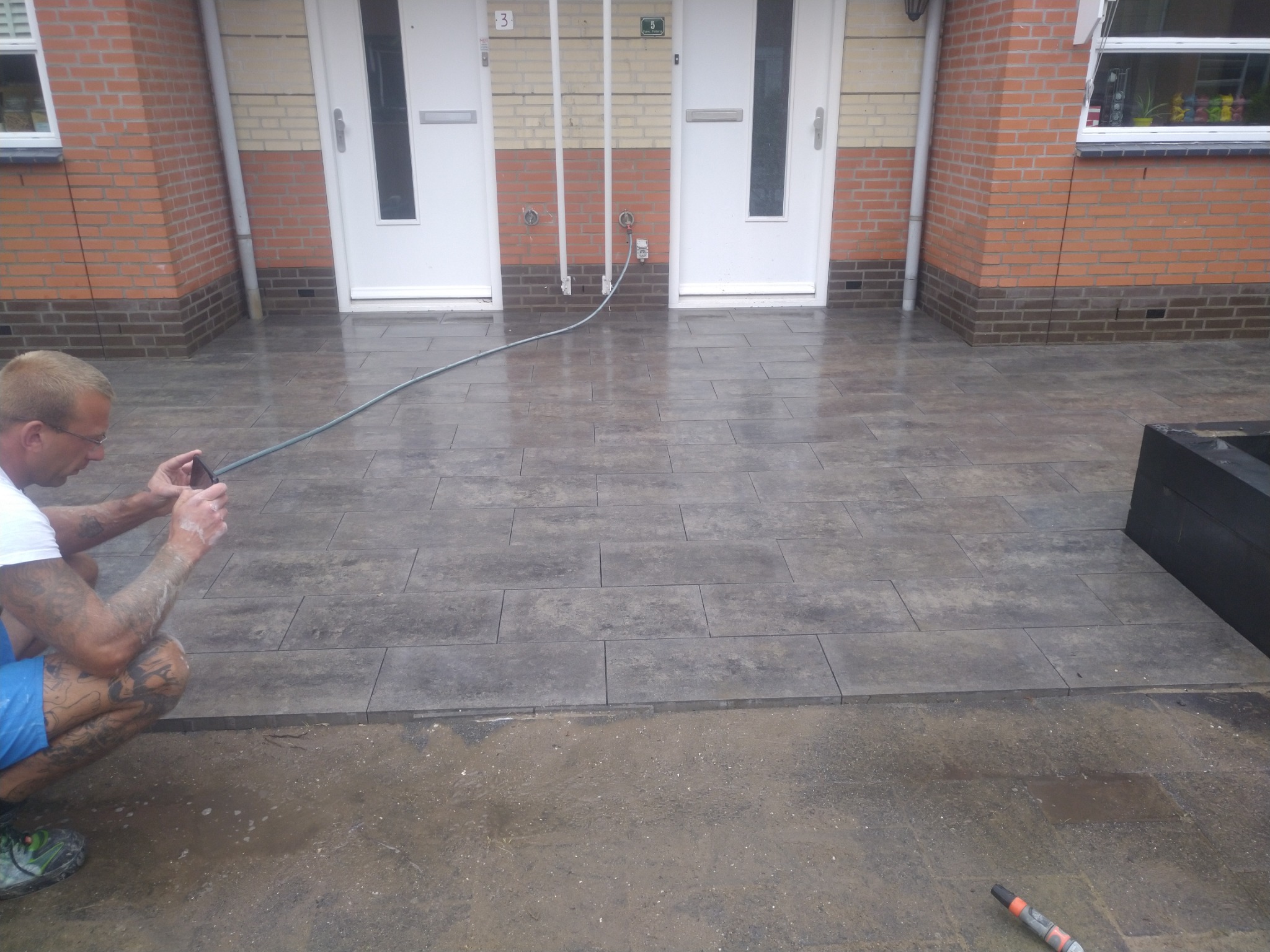 Ułożona szara kostka brukowa przed budynkiem, pracownik robi zdjęcie telefonem, widoczne ślady wilgoci i niedokończone prace przy krawędzi.