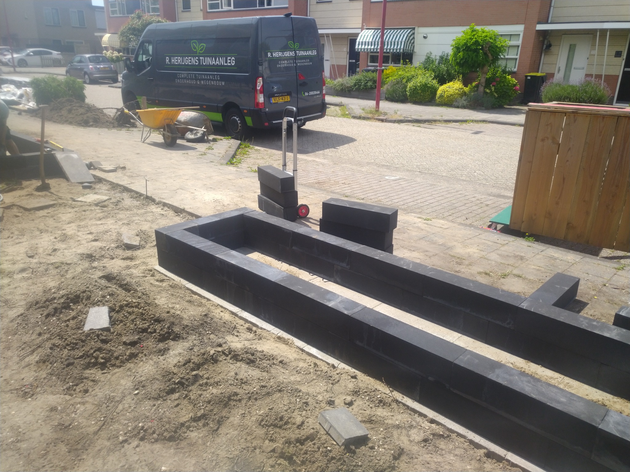 Przygotowanie podłoża pod taras z elementów betonowych, widoczny wózek transportowy, taczka i samochód firmy budowlanej w tle.