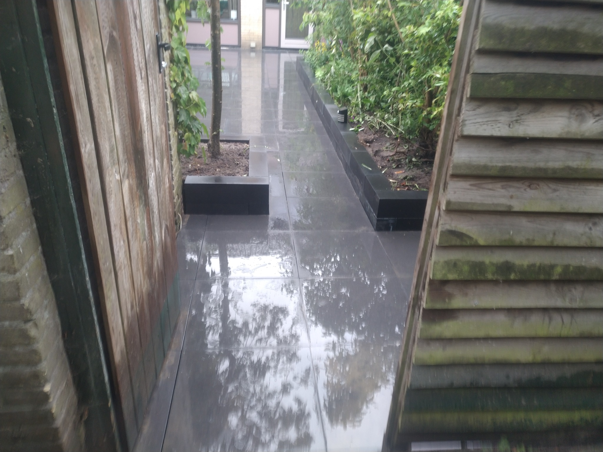 Śliska, mokra nawierzchnia z szarych płyt, otoczona drewnianym płotem i zielenią, odbijająca niebo.