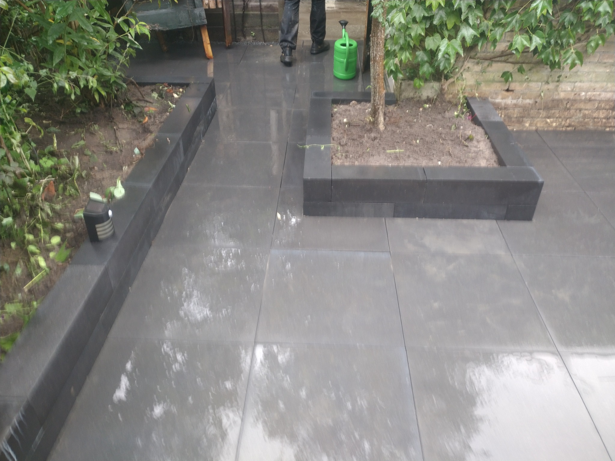 Świeżo ułożony, mokry taras z szarych płyt kamiennych z obrzeżami z ciemnego kamienia, obok zielony krzew i konewka