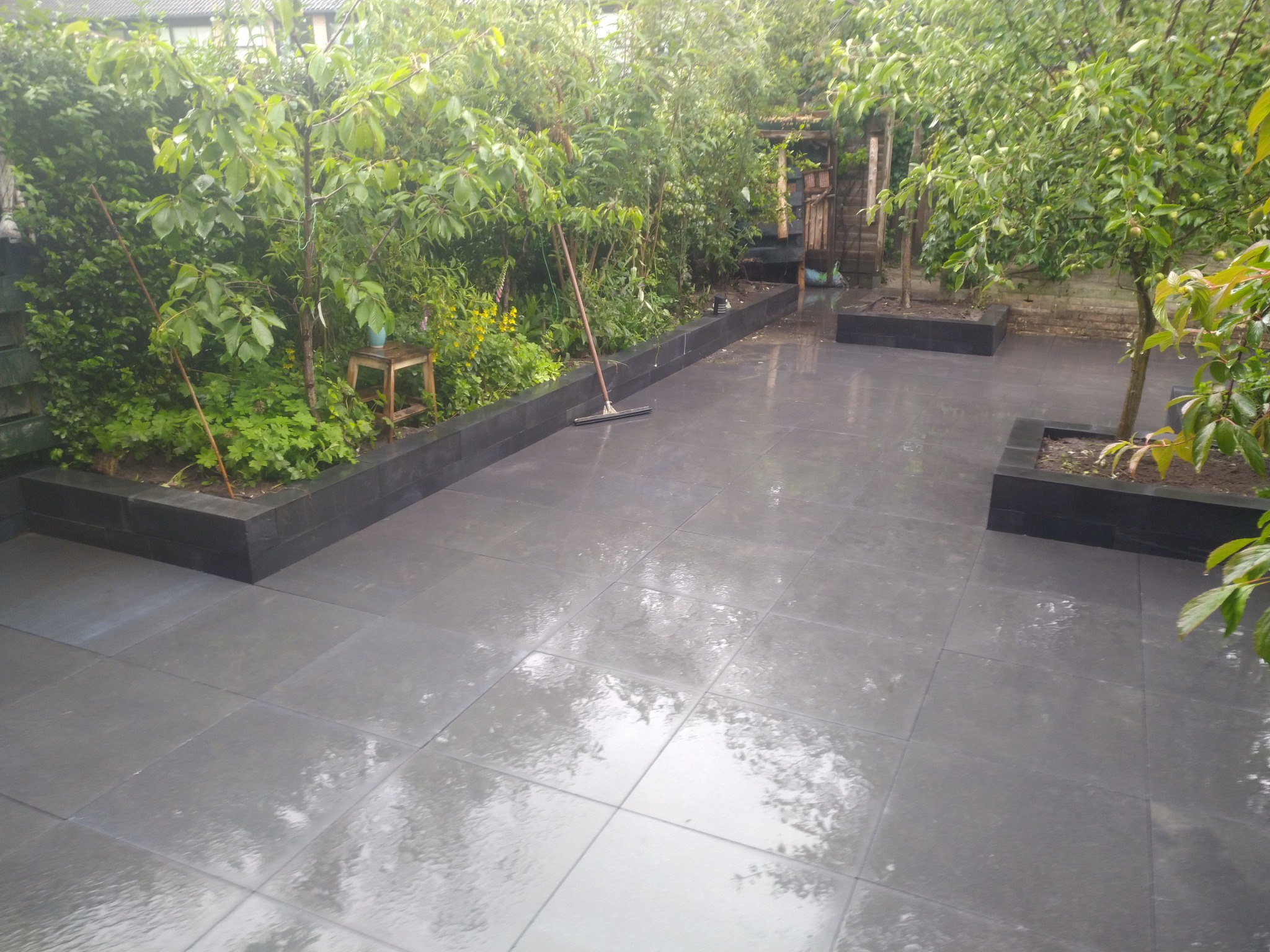 Szara, prostokątna nawierzchnia z płyt, otoczona zielenią i niskimi murkami, z widocznymi refleksami po deszczu.