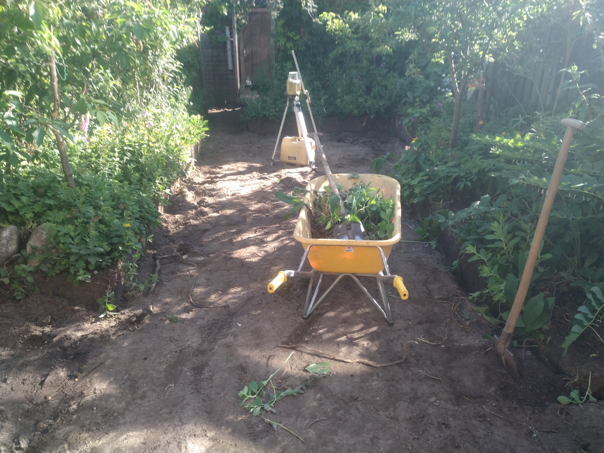 Przygotowanie terenu pod ścieżkę w ogrodzie: wykopany grunt, taczka z chwastami, niwelator laserowy i szpadel.
