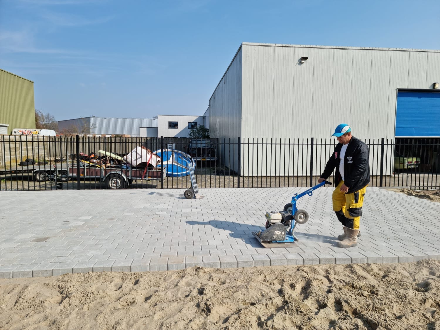 Pracownik zagęszcza wibroprasa ułożoną szarą kostkę brukową na posesji, widoczny piasek po bokach i ogrodzenie z metalowych prętów w tle.