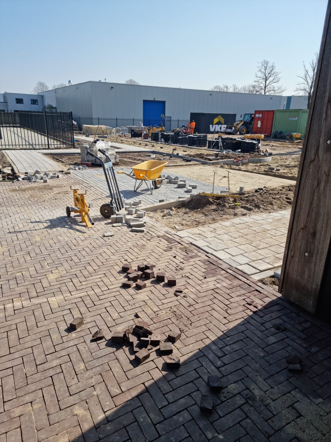 Plac budowy z częściowo ułożoną czerwoną kostką w jodełkę, widoczne narzędzia brukarskie, taczka i palety z kostką w tle, budynek magazynowy w oddali.