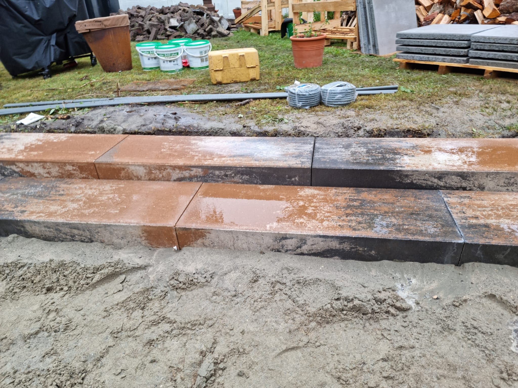 Dwa stopnie schodów zewnętrznych z kamiennych bloków o zróżnicowanej kolorystyce, częściowo mokre od deszczu, osadzone w piasku; w tle widoczne materiały budowlane i narzędzia.