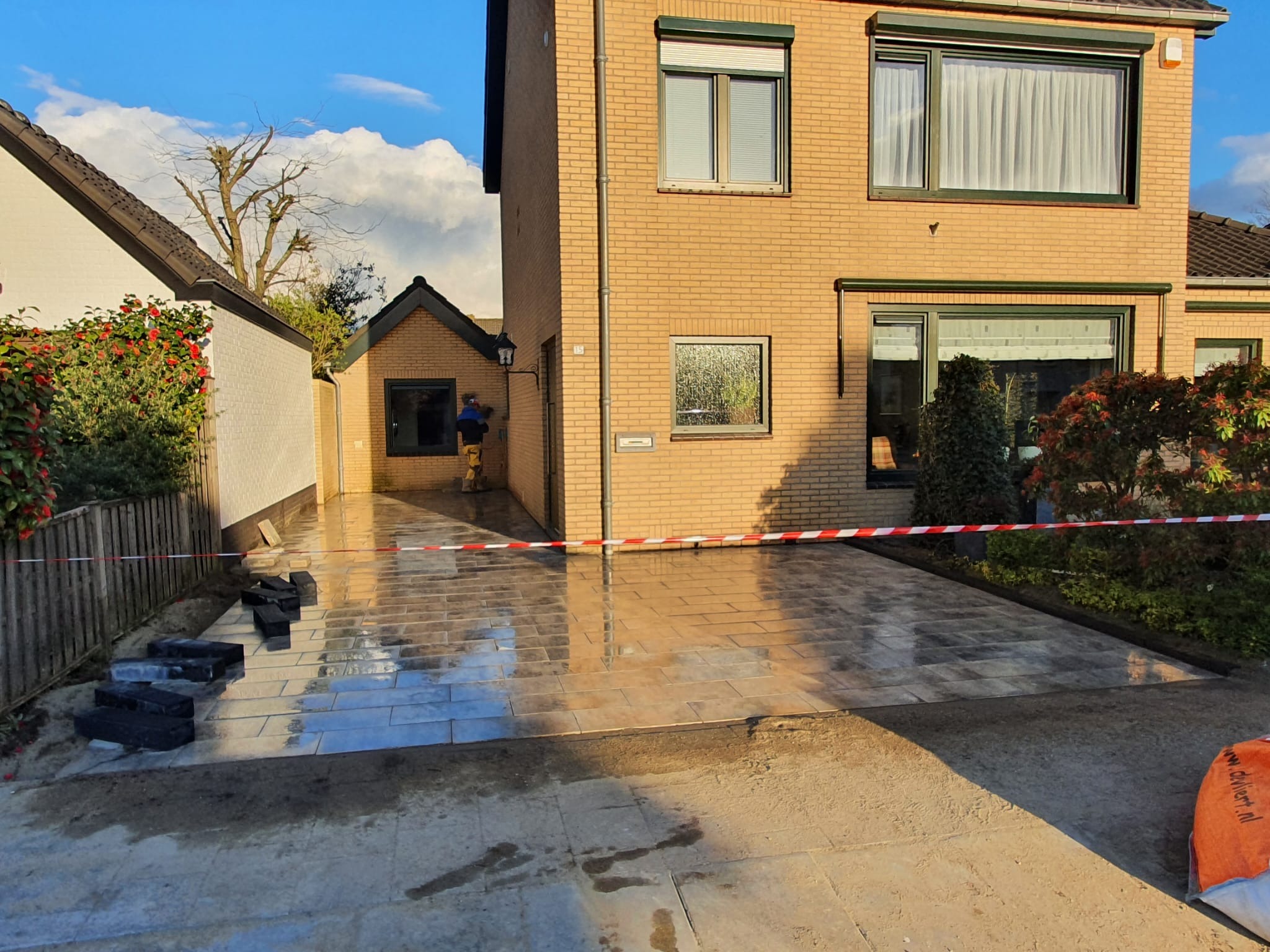 Świeżo ułożona, mokra nawierzchnia z prostokątnej kostki brukowej w odcieniach szarości, częściowo zabezpieczona taśmą ostrzegawczą, z widocznym pracownikiem w oddali i stosem kostki do ułożenia...