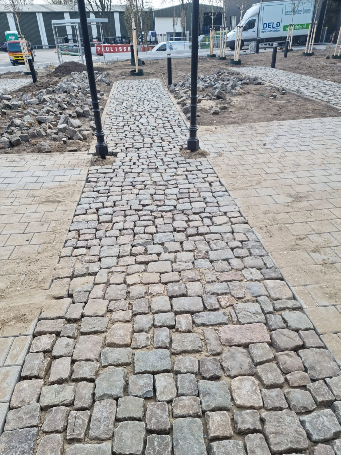 Ścieżka z kostki granitowej w trakcie układania, widoczne czarne lampy, piasek i elementy kostki do ułożenia.