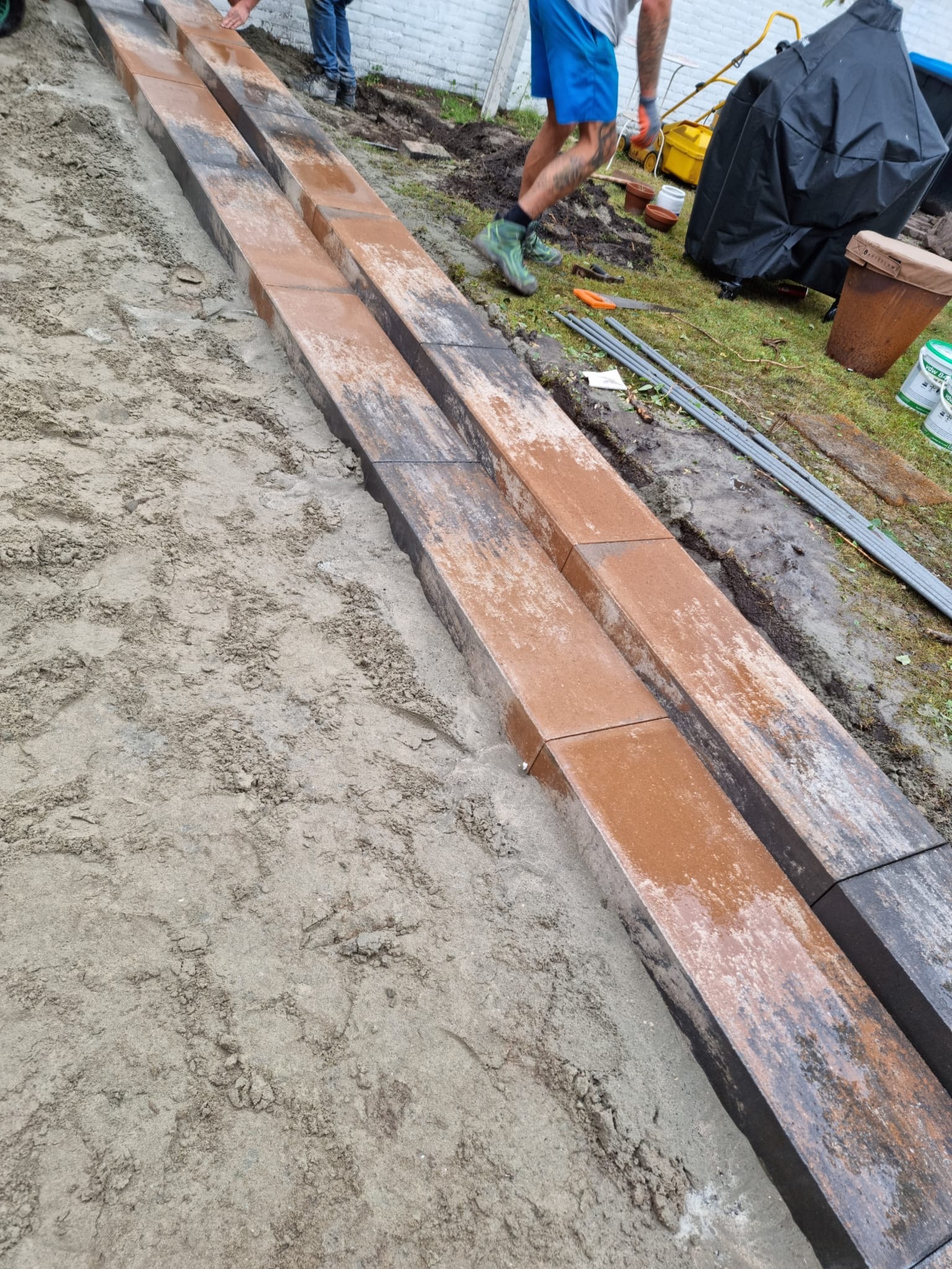 Montaż obrzeży chodnika z kostki brukowej w piaskowym podłożu, widoczne narzędzia i fragment trawnika w tle.