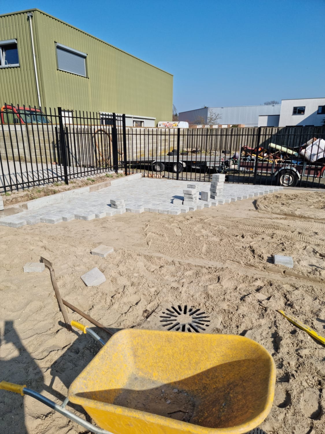 Fragment układanej nawierzchni z szarej kostki brukowej na piasku, widoczna żółta taczka, łopata i poziomica, czarne ogrodzenie w tle, budynek magazynowy.
