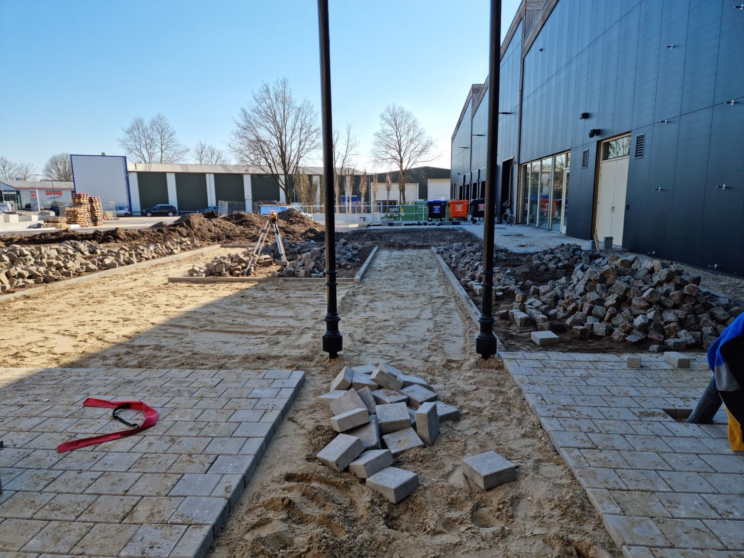 Plac budowy z częściowo ułożoną szarą kostką brukową, widoczne przygotowane podłoże z piasku, stosy kostki i narzędzia pomiarowe w tle, budynek z czarną elewacją.