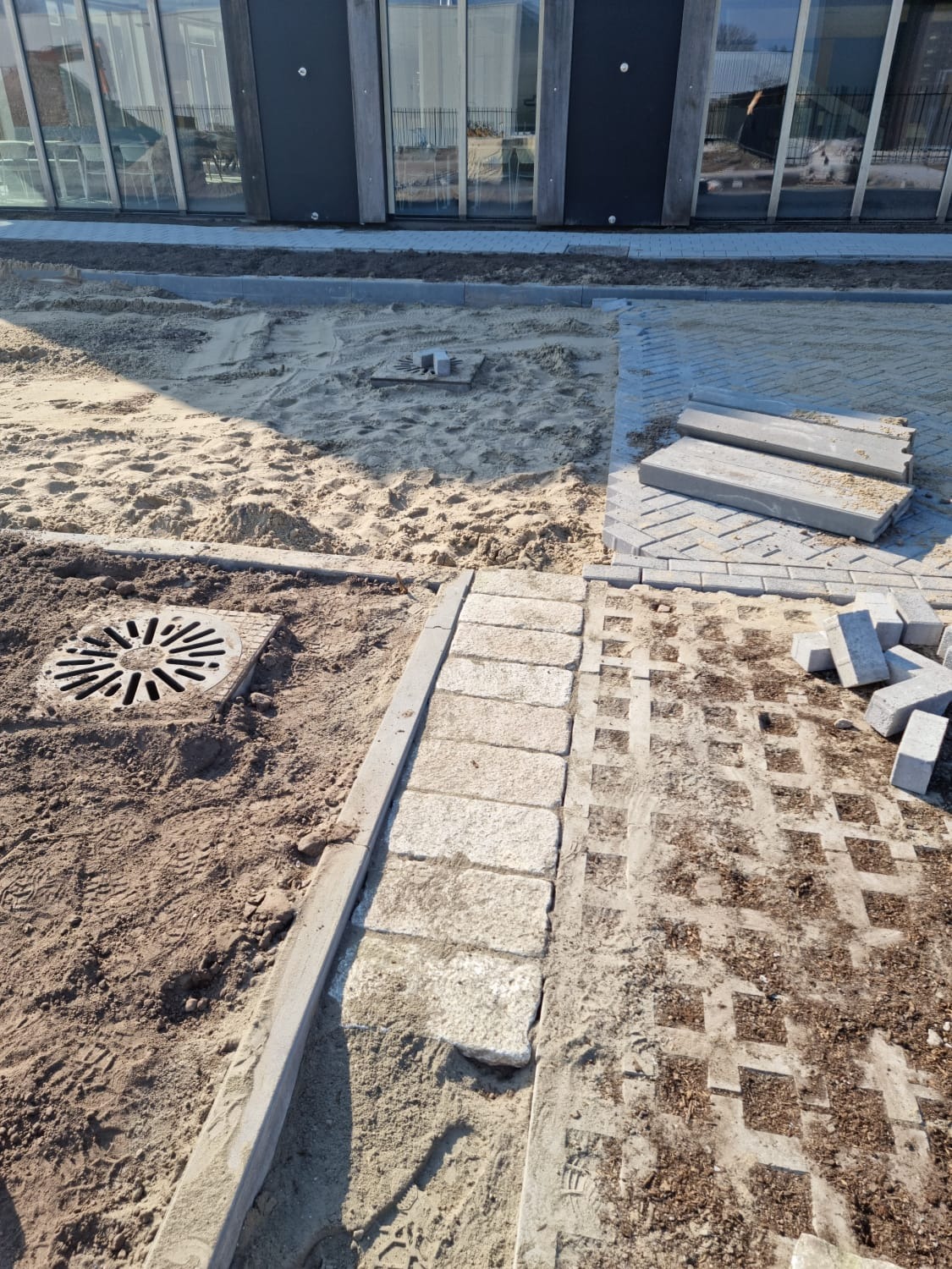 Etap prac brukarskich: przygotowanie podłoża z piasku, ułożone obrzeża z kostki granitowej i fragmenty nawierzchni z kostki ażurowej, widoczny wpust kanalizacyjny.