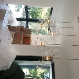 Nowoczesny żyrandol z kulistymi kloszami zamontowany w jasnym wnętrzu, widoczne okna z widokiem na zieleń, częściowo zakryte kartony i folia ochronna sugerujące trwający remont.