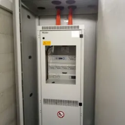 Otwarta szafa elektryczna z widocznymi przewodami wchodzącymi od góry, tablicami kontrolnymi i ostrzeżeniami o napięciu.