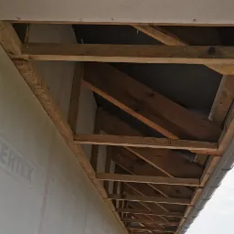 Surowa konstrukcja drewniana dachu wystająca poza ścianę domu w trakcie budowy, widoczne elementy ocieplenia i rynna, w tle drugi budynek i stos materiałów budowlanych.