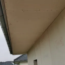 Montaż podbitki dachowej z widocznymi elementami mocującymi i rynnami, na tle ocieplonej ściany budynku z widocznym fragmentem rusztowania.