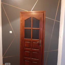 Szara ściana z geometrycznym wzorem z cienkich, złotych linii, drewniane drzwi z szybą w centrum kadru, widoczny włącznik światła.