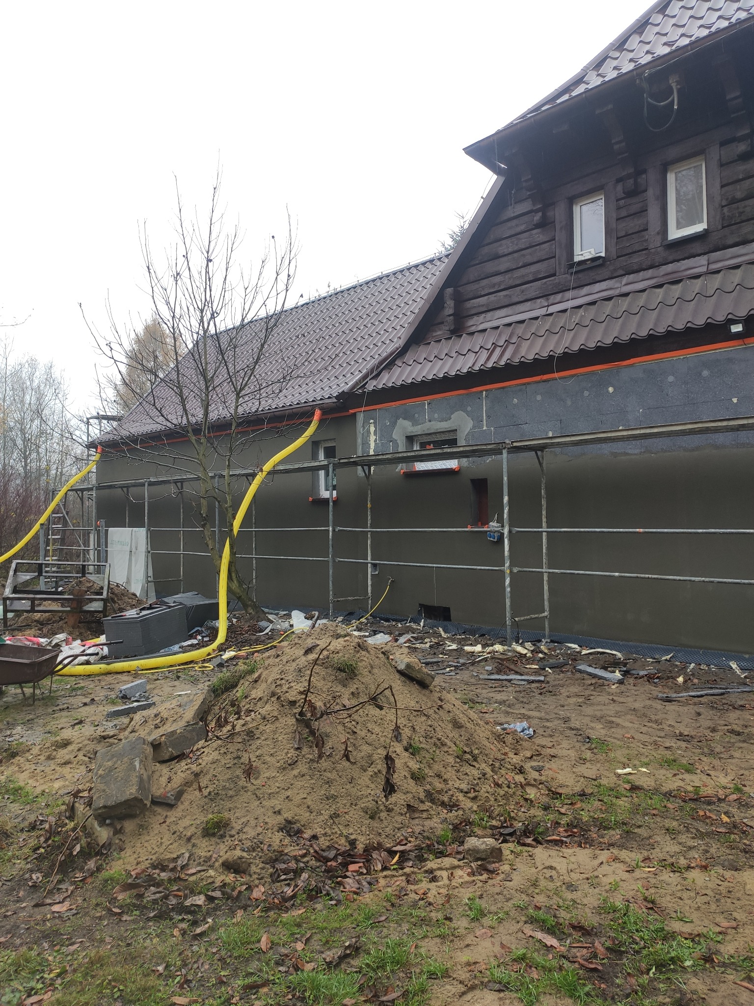 Dom z drewnianą górą i elewacją w trakcie ocieplania, widoczne rusztowania, żółte rury wentylacyjne i sterta piasku na pierwszym planie.