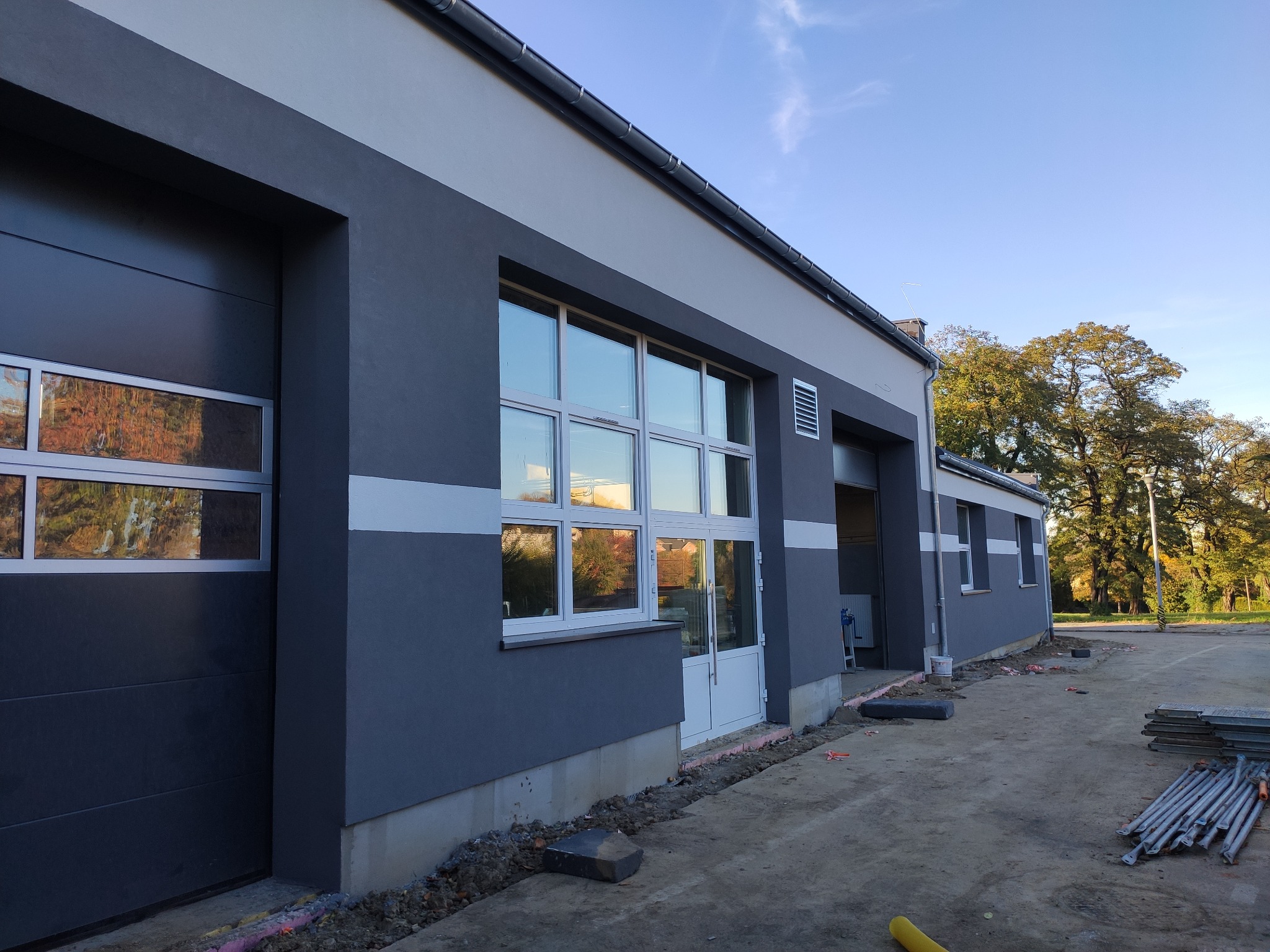 Nowoczesny budynek w odcieniach szarości z białymi akcentami, widoczne okna z refleksami drzew, teren wokół budynku w trakcie prac wykończeniowych, widoczne materiały budowlane.